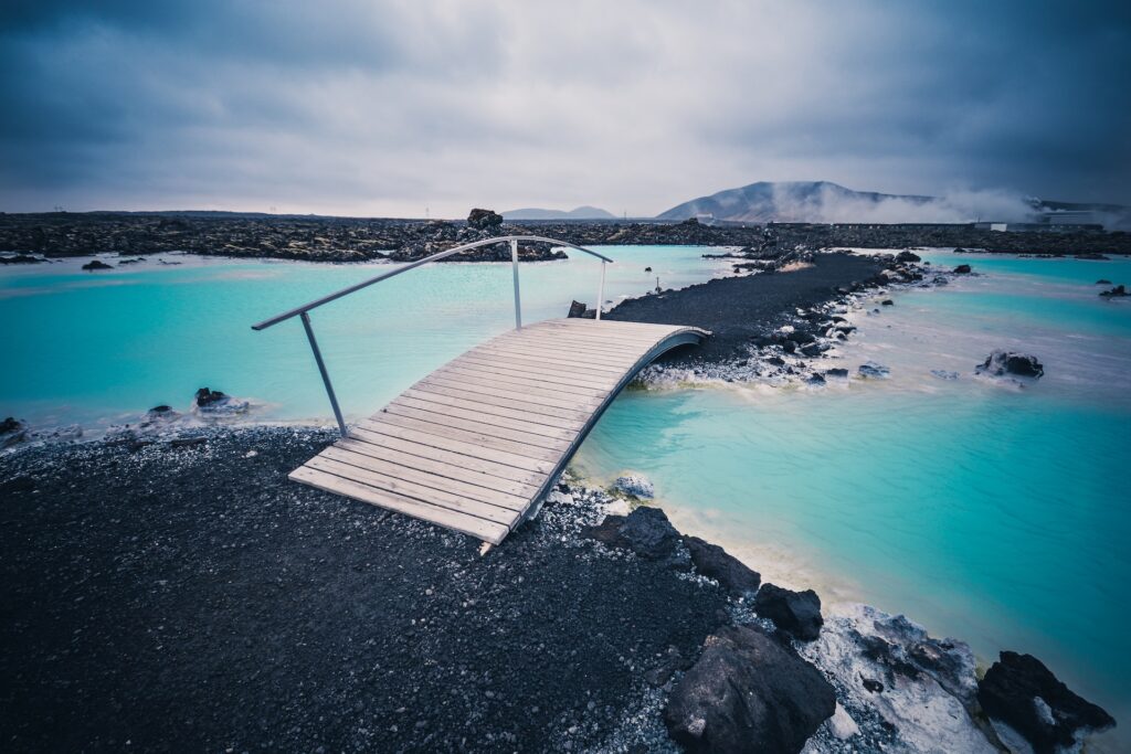 Daytime picture showing the blue lagoon with a foot bridge over a small section of water and rocks round the edge of the lagoon